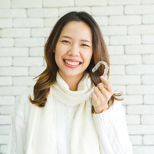 A young female wearing a white sweater and cream-colored scarf holds an Invisalign aligner in her left hand