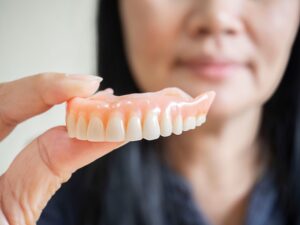 Woman blurry in background holding dentures in foreground with two fingers